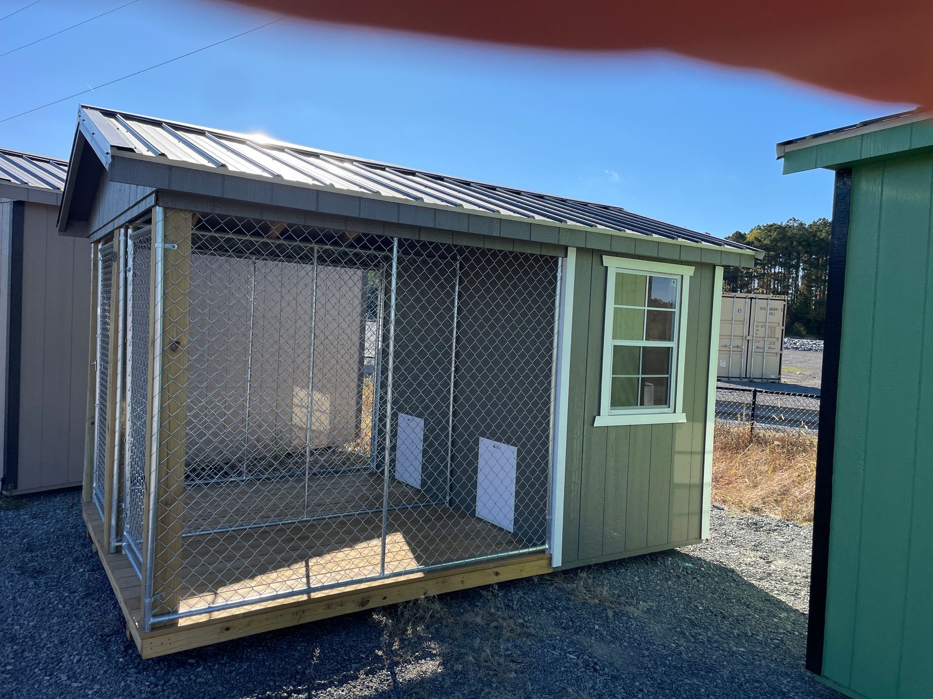 A small, sage-green wooden outdoor kennel with a metal roof, chain-link front, and a single window.