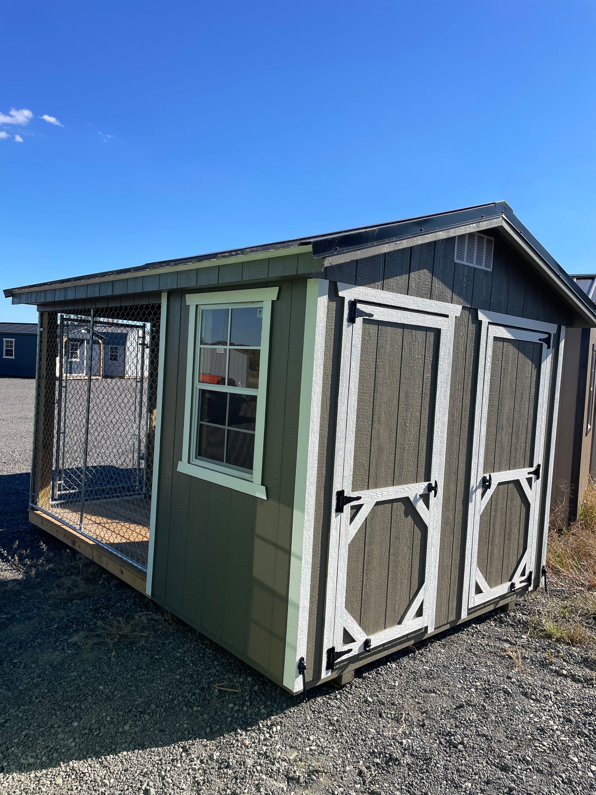 A green and brown outdoor shed with white-trimmed double doors and a window, sitting on a gravel lot under a blue sky.
