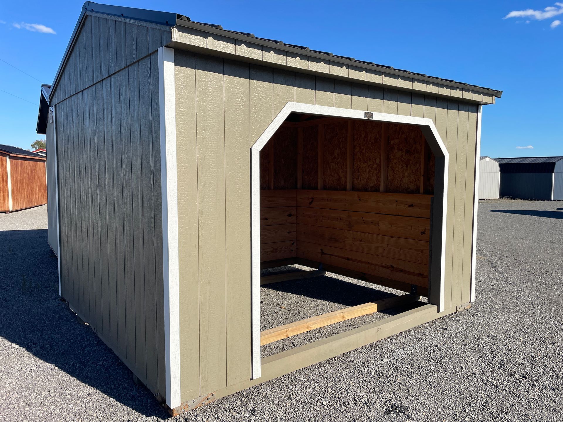 Tan run-in shed with white trim and an open front, sitting on a gravel lot under a clear blue sky.
