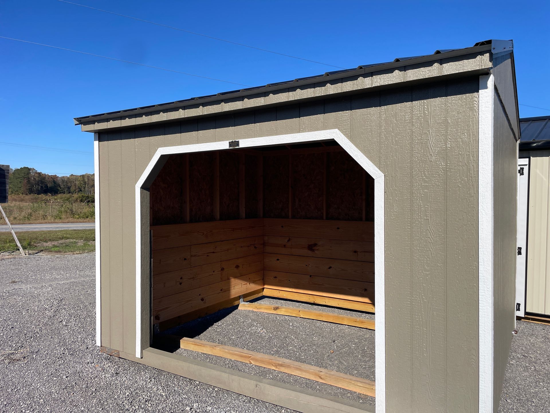 A tan-colored wooden run-in shed with a metal roof, white trim, and interior wooden board walls sitting on a gravel lot.
