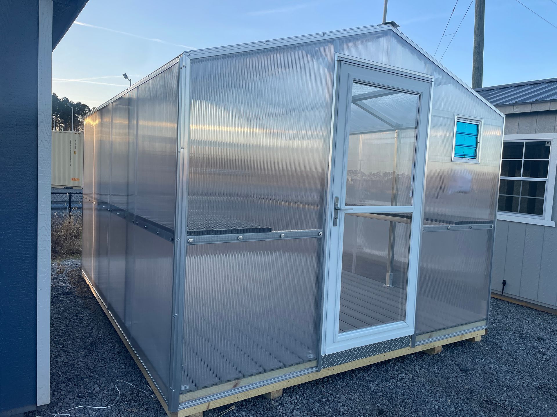A small, modern greenhouse with translucent ribbed walls, a white-framed door, and a wooden floor, set on a gravel lot.
