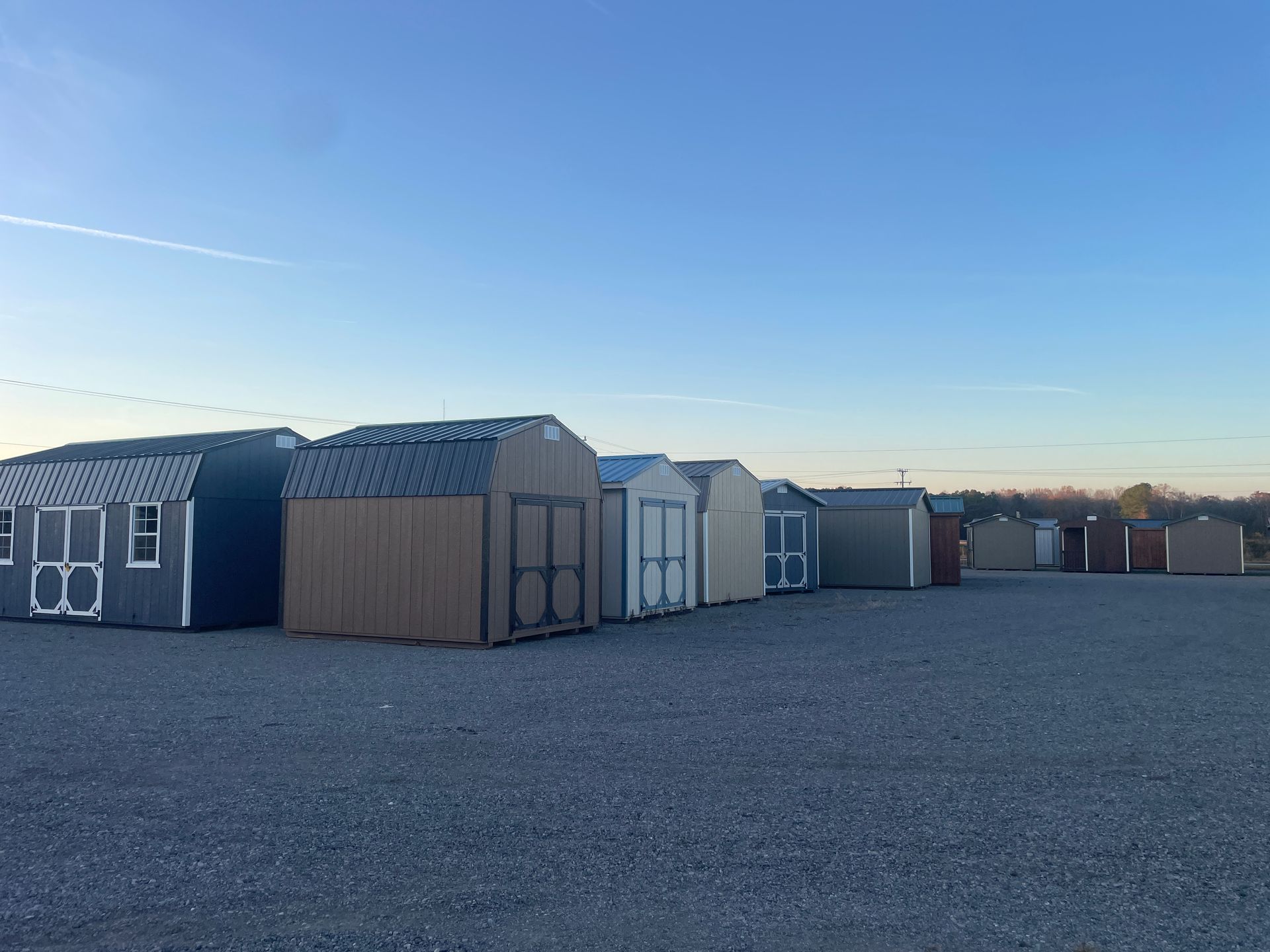 A row of various prefabricated storage sheds on a gravel lot under a clear blue sky.