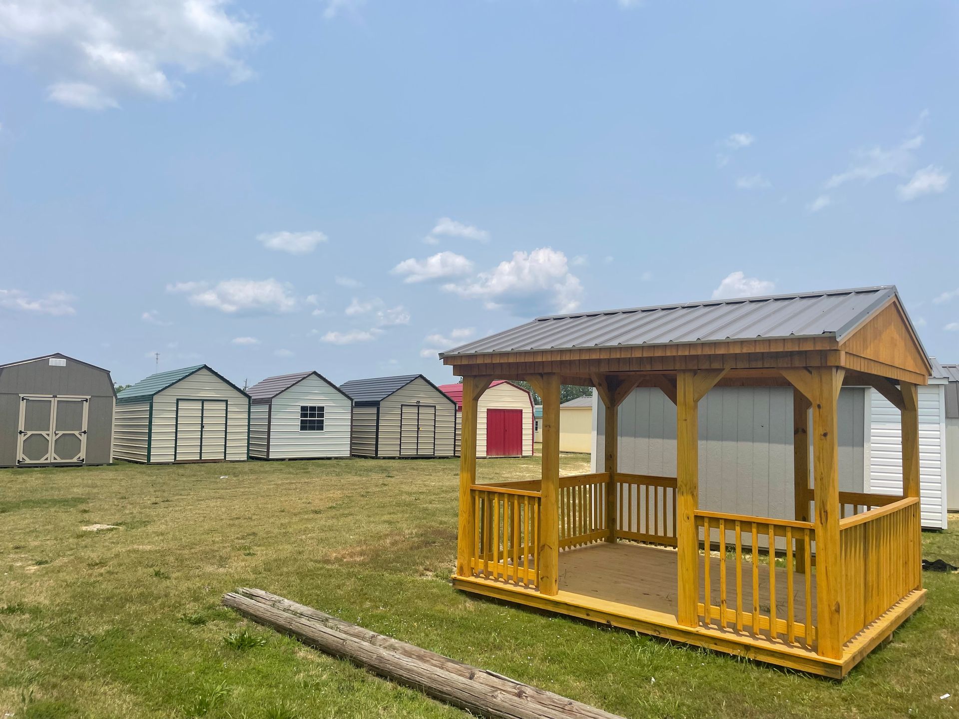 A wooden gazebo stands in a field with several various sheds in the background under a blue, partly cloudy sky.