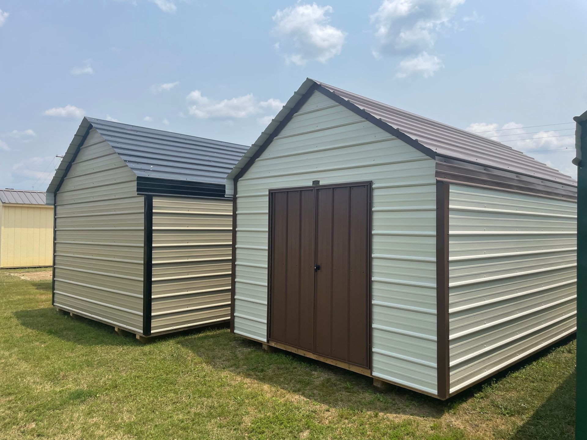 Two metal storage sheds with beige siding and dark brown trim sit on a grassy lot under a blue sky.