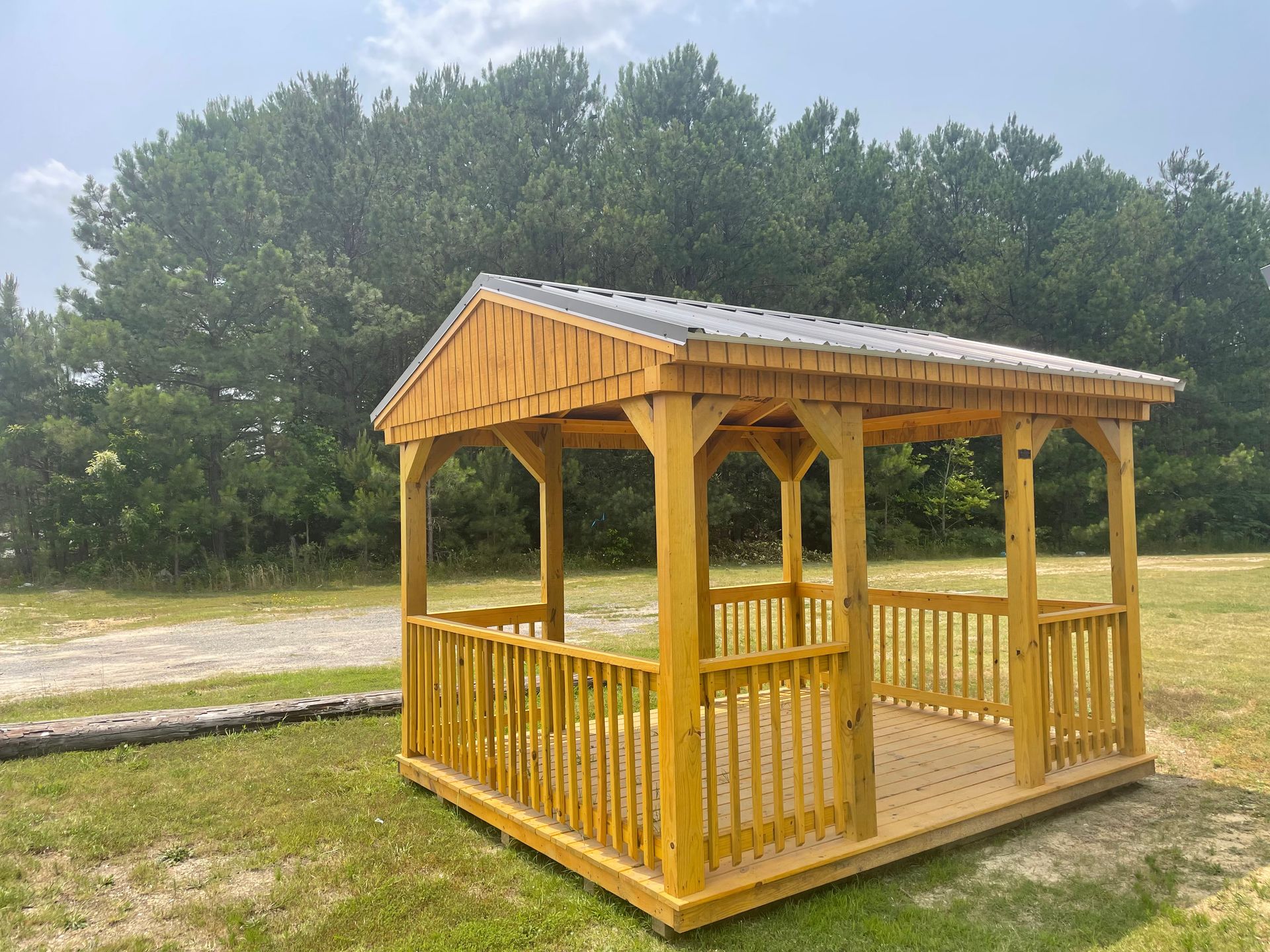 A wooden pavilion with a metal roof and railing, situated on a grassy field in front of a line of trees.