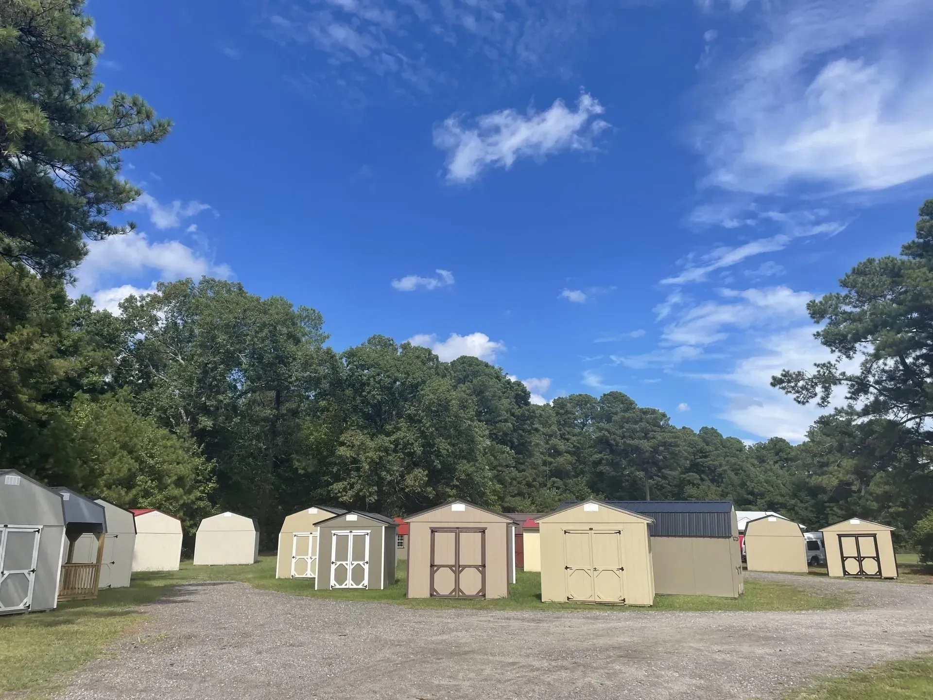 A collection of various storage sheds arranged on a gravel lot under a clear blue sky, surrounded by dense trees.