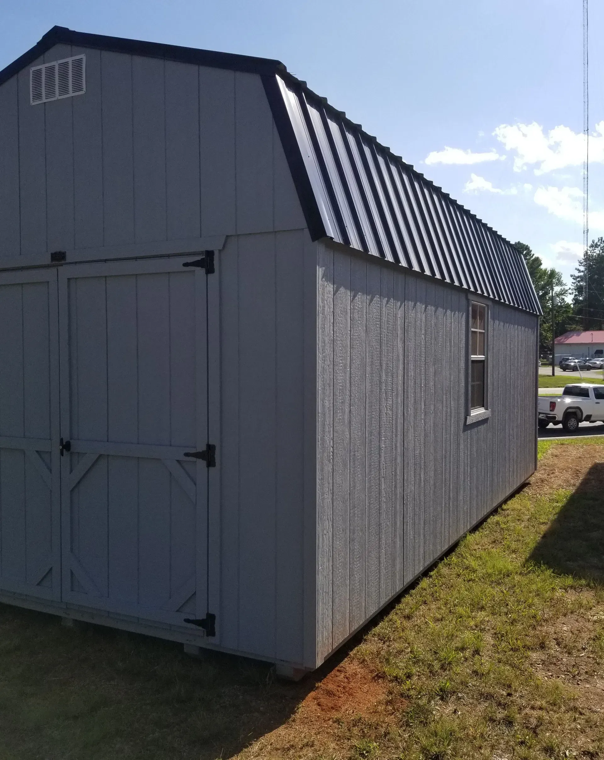 A grey gambrel-roof shed with a black metal roof and double doors, standing in a grassy field on a sunny day.