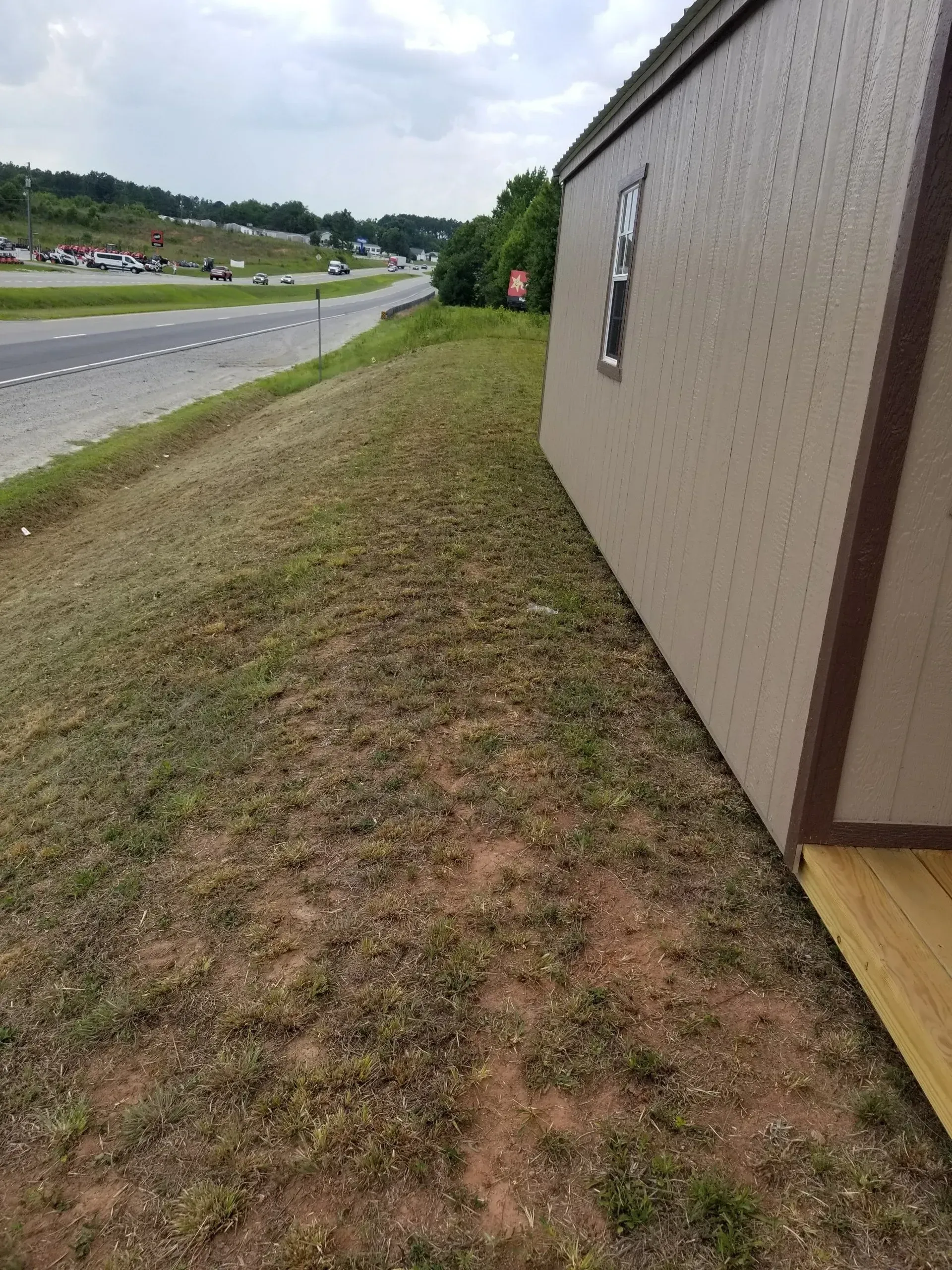 A view along the side of a tan shed on a grassy embankment next to a road under a cloudy sky.