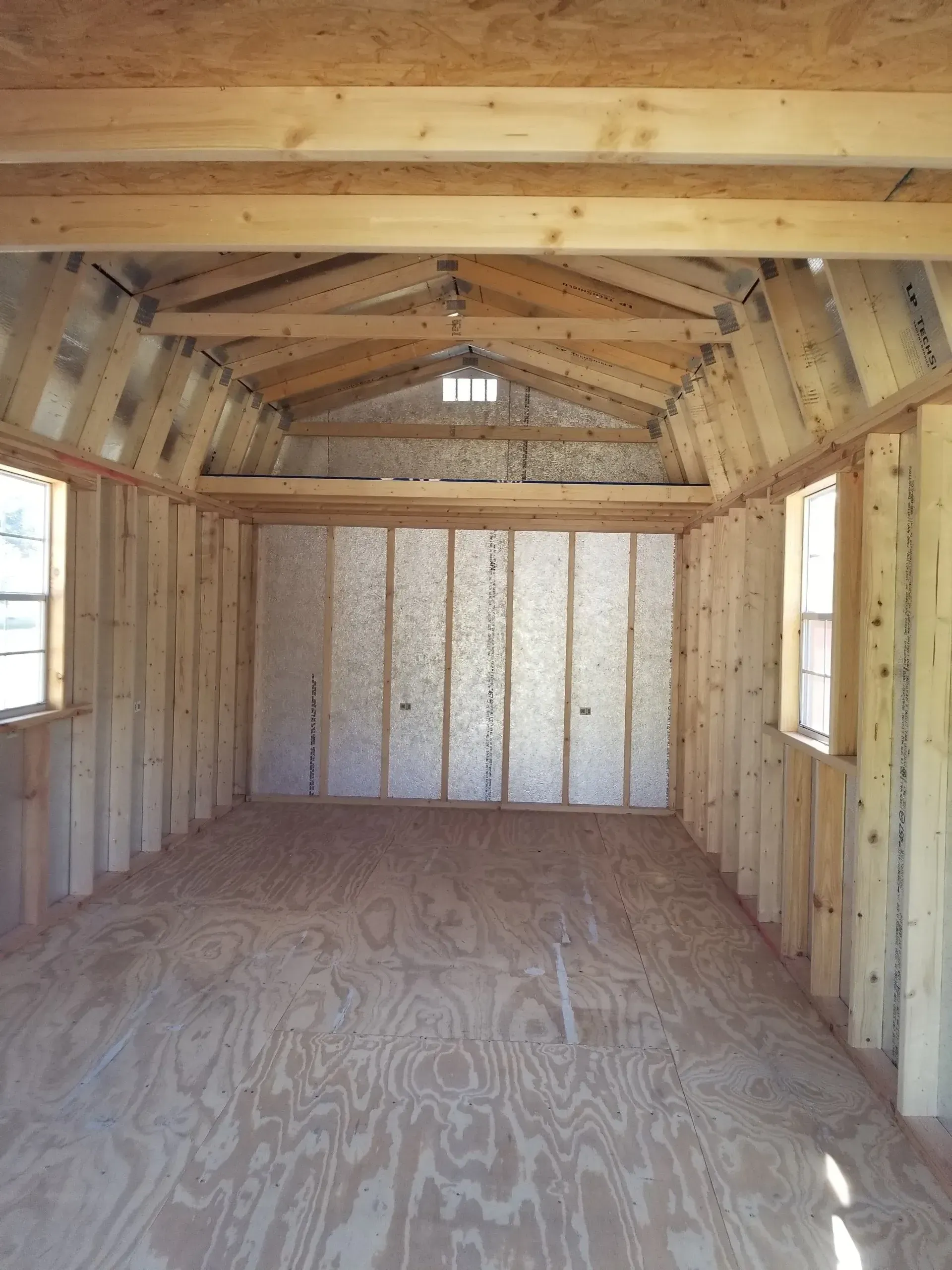 An interior view of a wooden shed with exposed studs, plywood flooring, windows on both sides, and a small loft area.