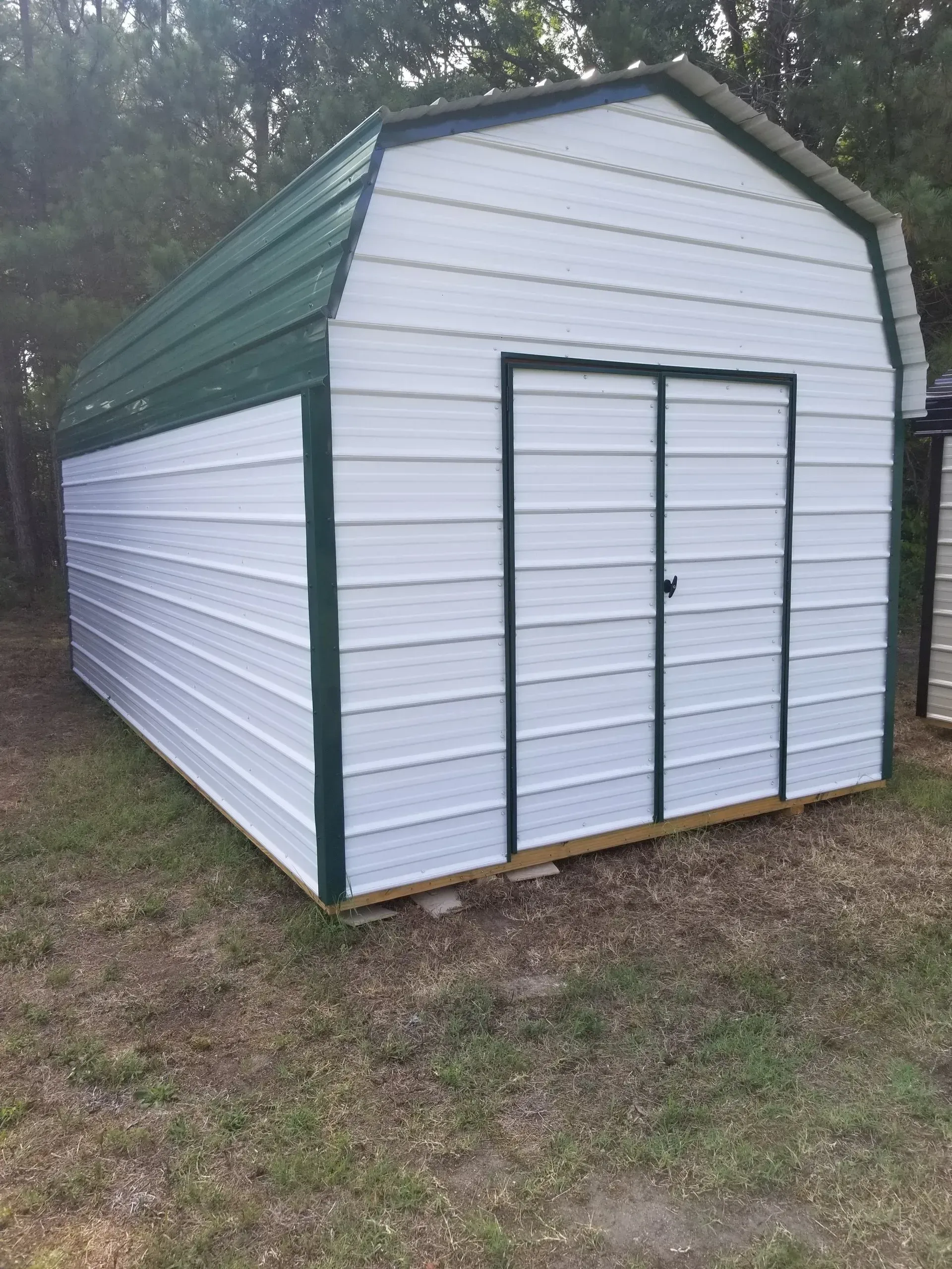 A white metal storage shed with a green roof and trim, sitting on grass in a wooded area.
