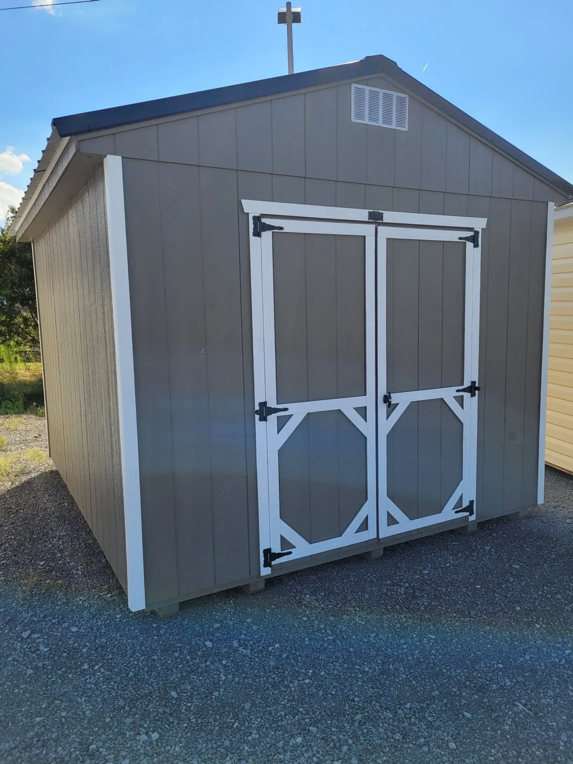 A grey, wooden storage shed with white trim and double doors, standing on a gravel surface under a clear blue sky.