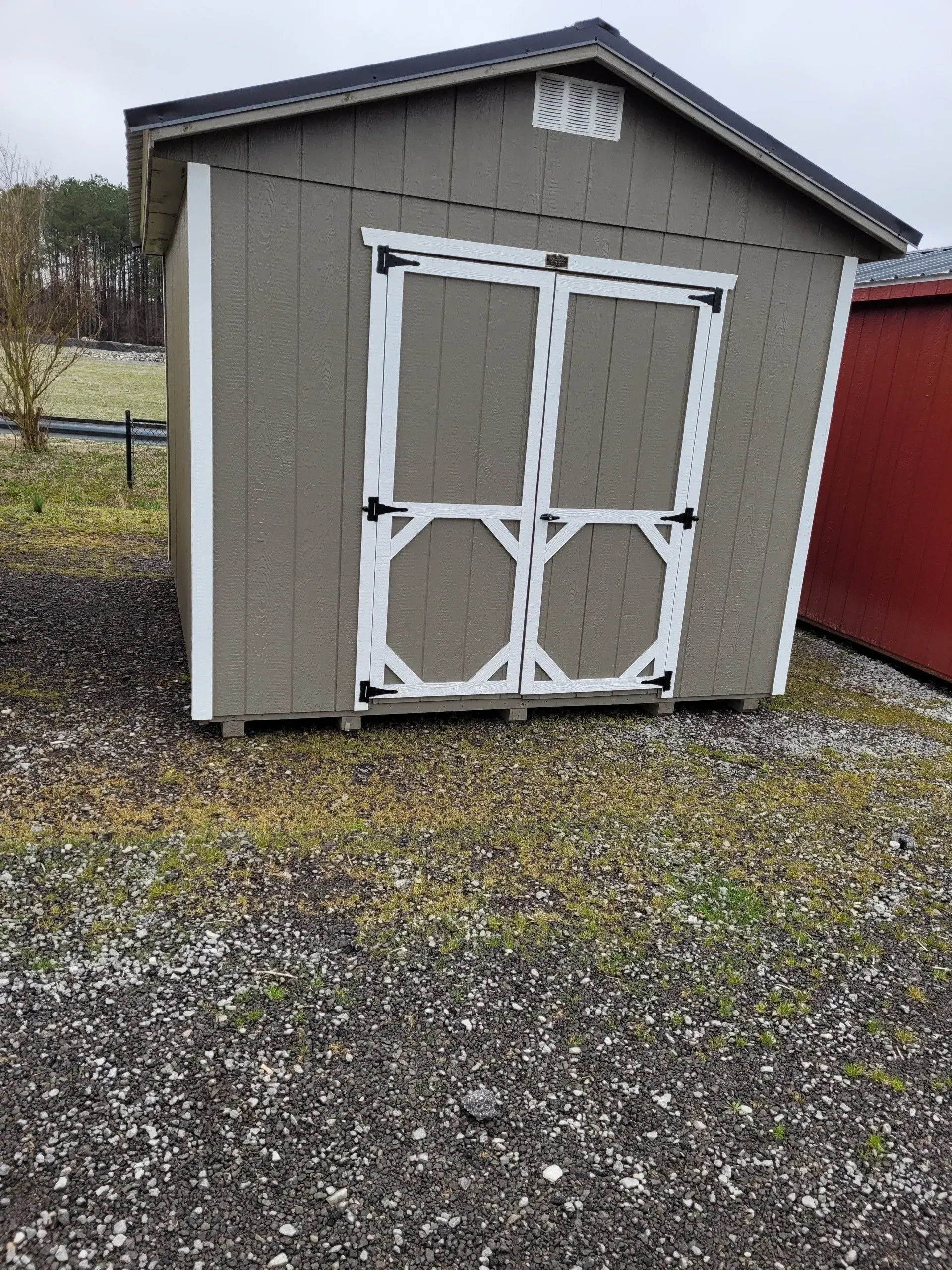 A taupe storage shed with white double doors and trim, situated on a gravel surface outdoors under an overcast sky.