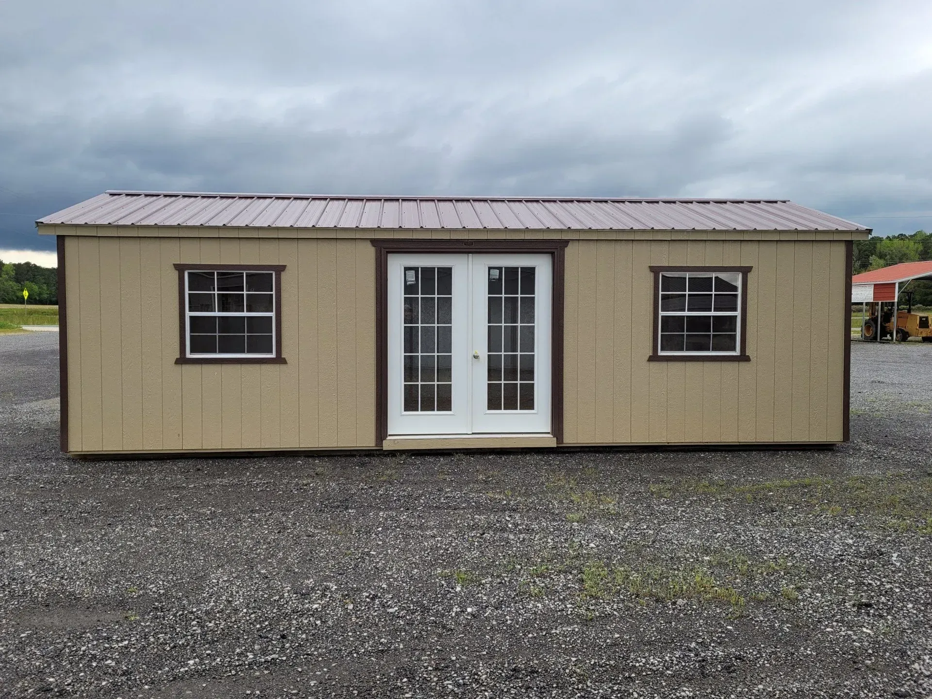 A tan-colored rectangular shed with a dark roof, two white double doors, and two windows, set on a gravel lot.