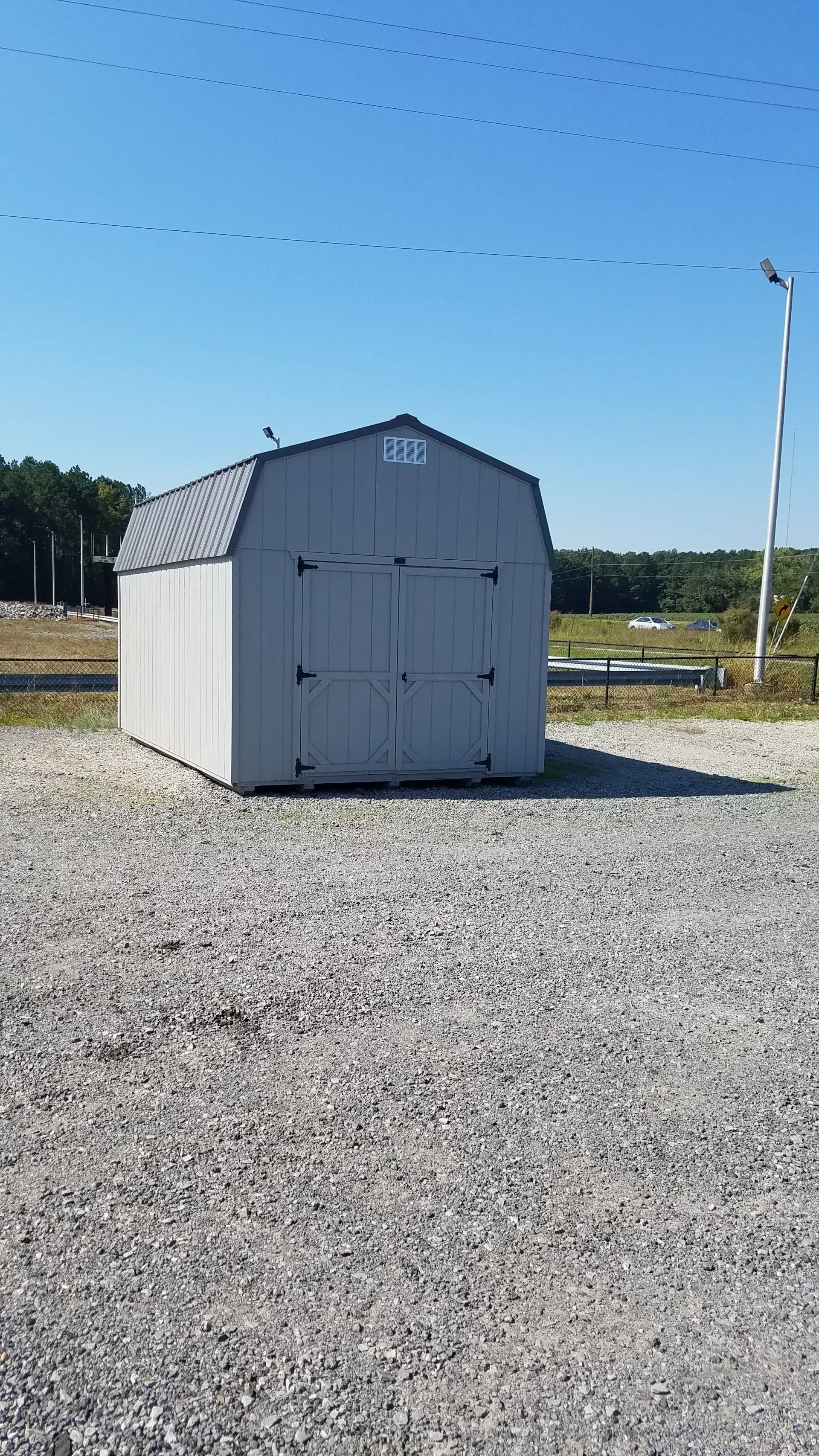 A gray, gambrel-roofed storage shed stands alone on a gravel lot under a clear blue sky.