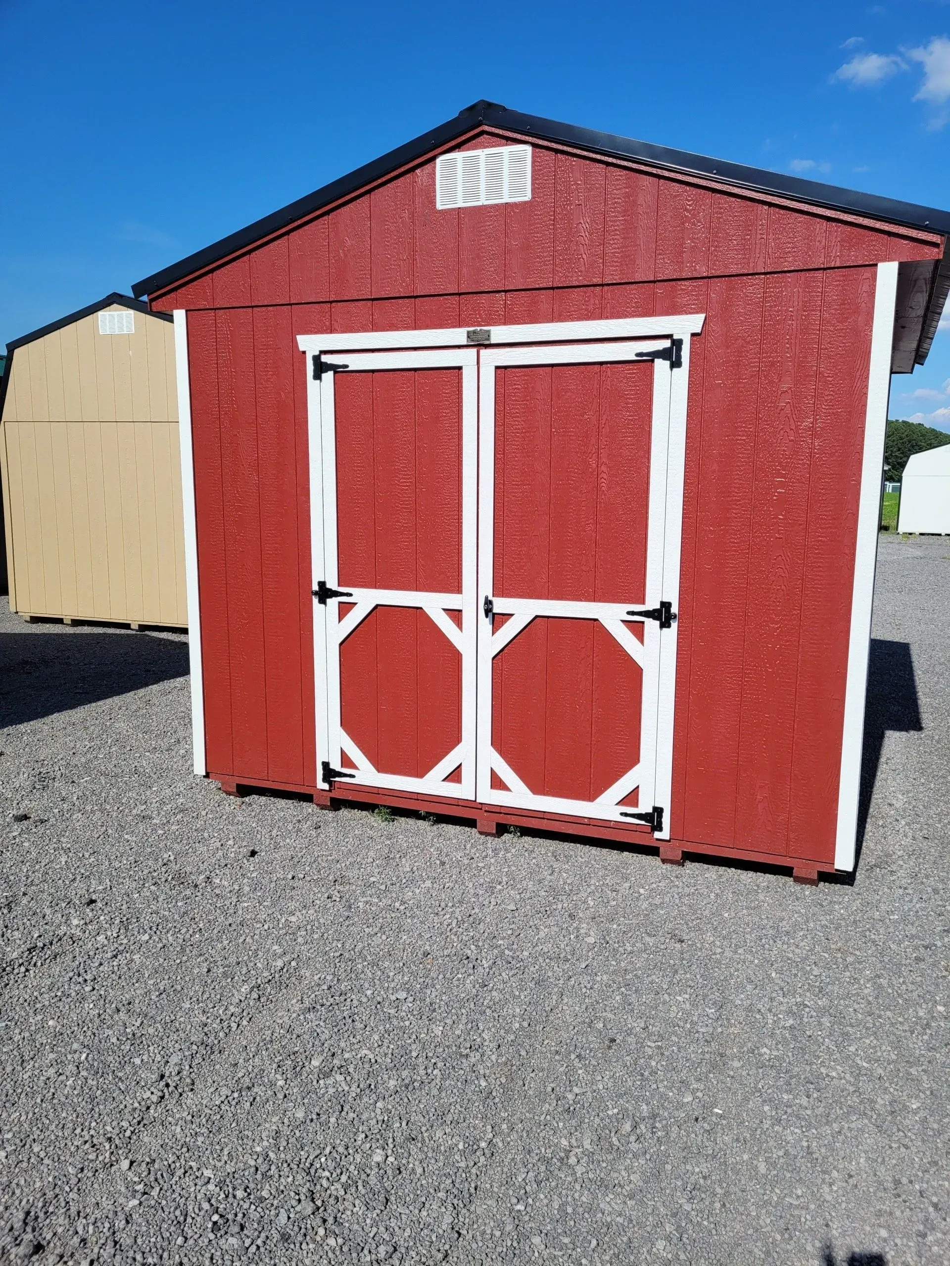 A red storage shed with white trim and double doors, sitting on a gravel lot under a bright blue sky.