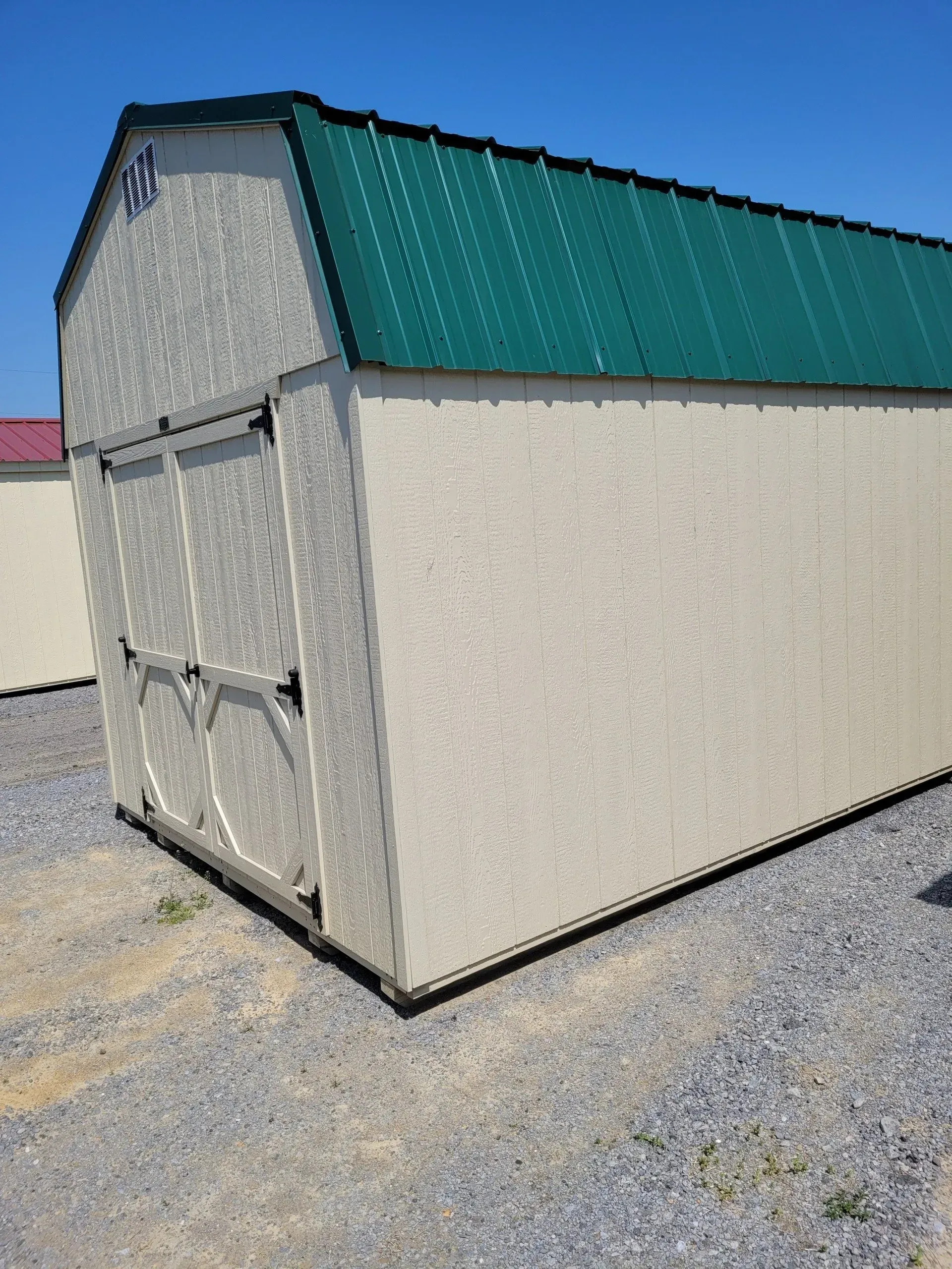 A tan outdoor storage shed with a green metal roof and double doors, standing on a gravel lot under a clear blue sky.