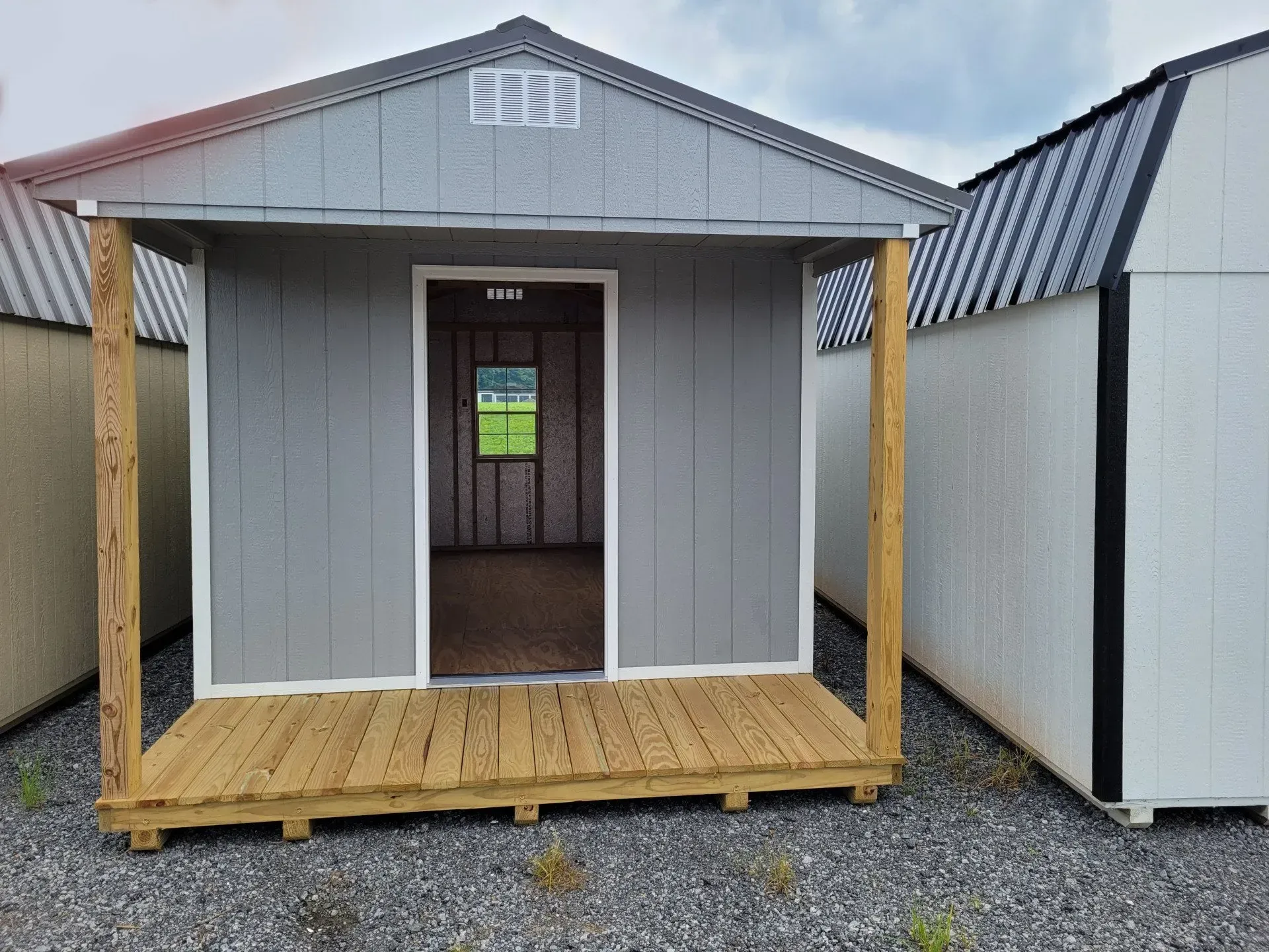 A grey wooden shed with an open door, a small porch, and a window visible inside, situated on a gravel lot.