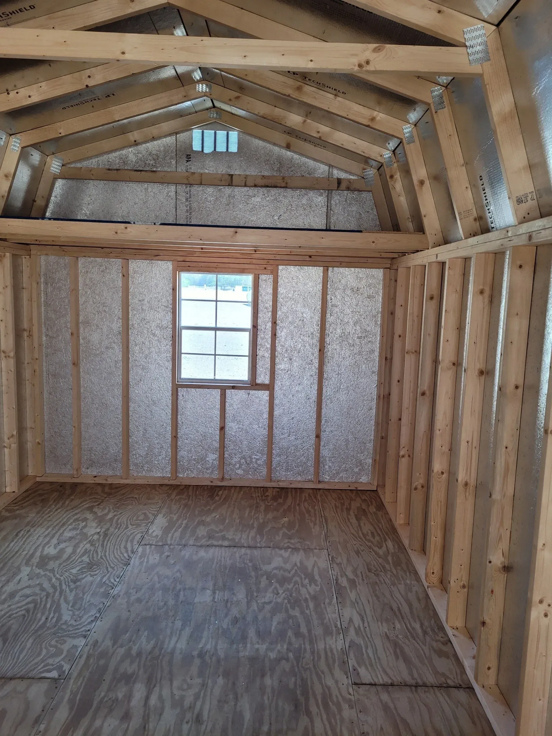 Interior view of an unfinished wooden storage shed with radiant barrier wall panels, a loft area, and one window.