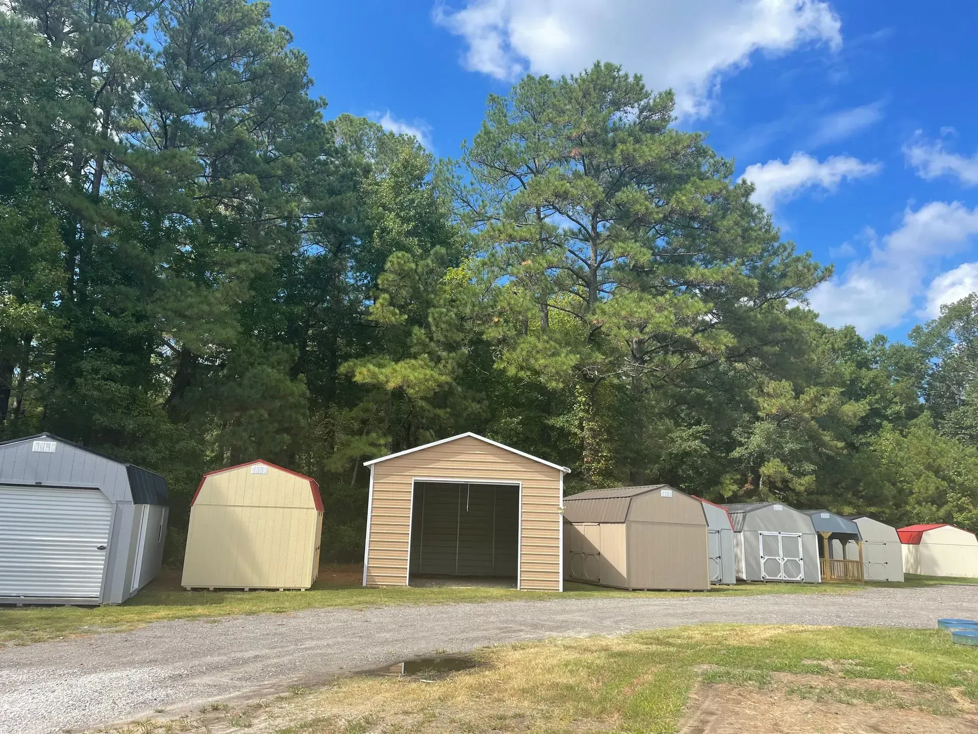 A row of various sheds and storage structures lined up on a gravel lot in front of a dense forest under a blue sky.