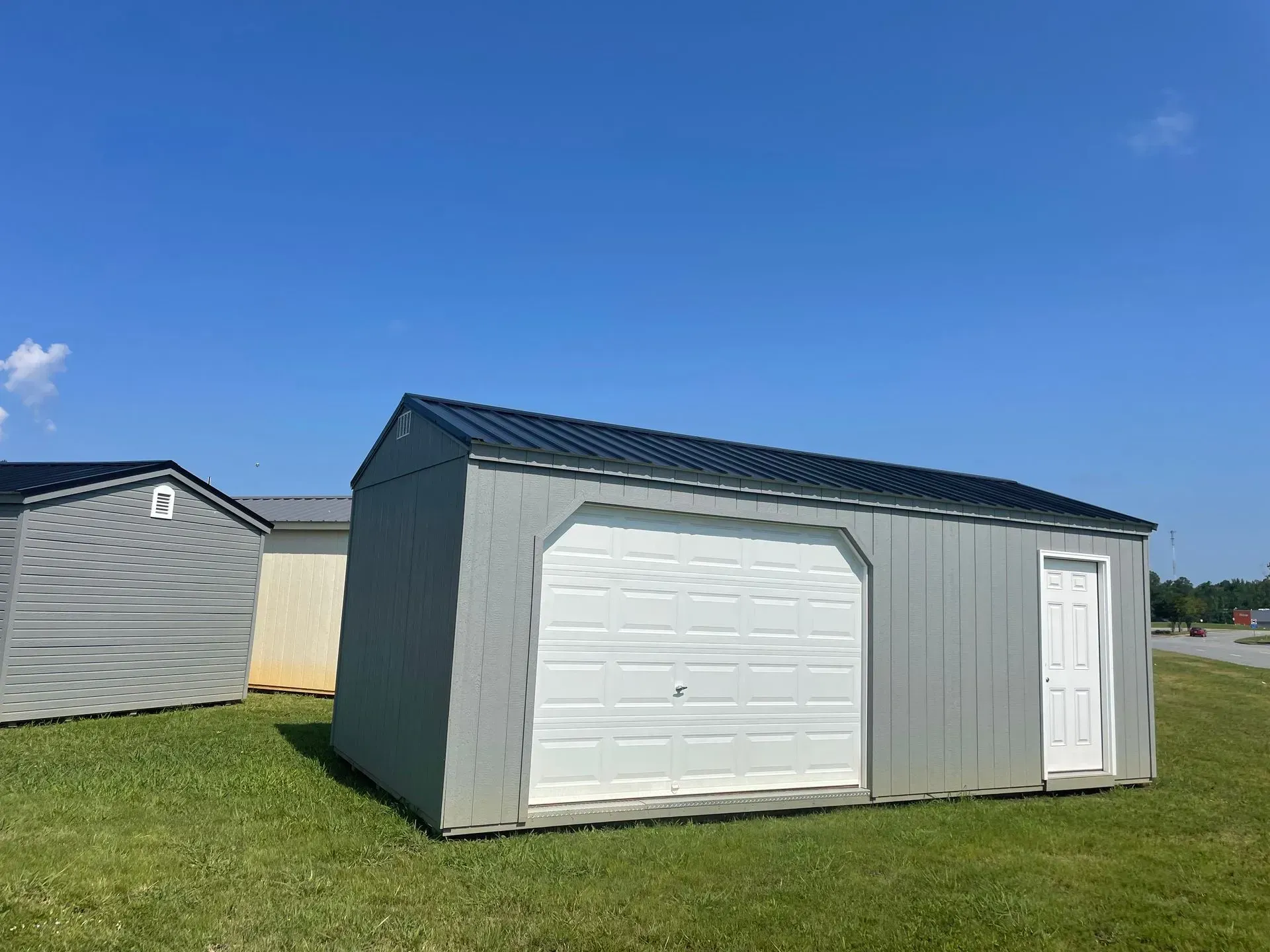 A grey metal shed with a white garage door and a side entry door, situated on a grassy lot under a clear blue sky.