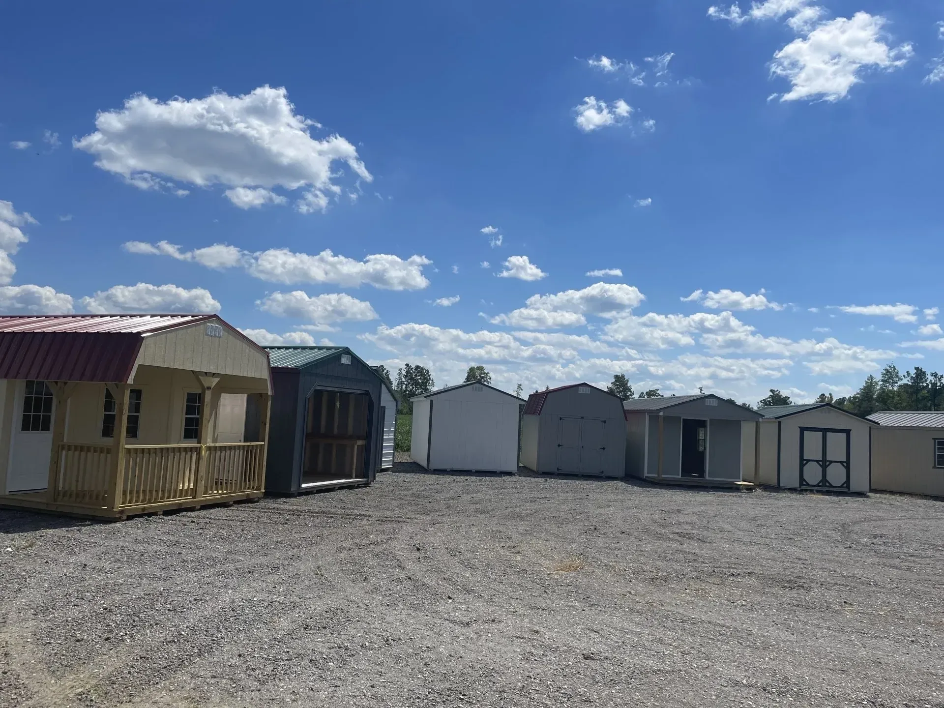 A gravel lot displays a row of various small pre-fabricated storage sheds under a bright blue sky with scattered clouds.