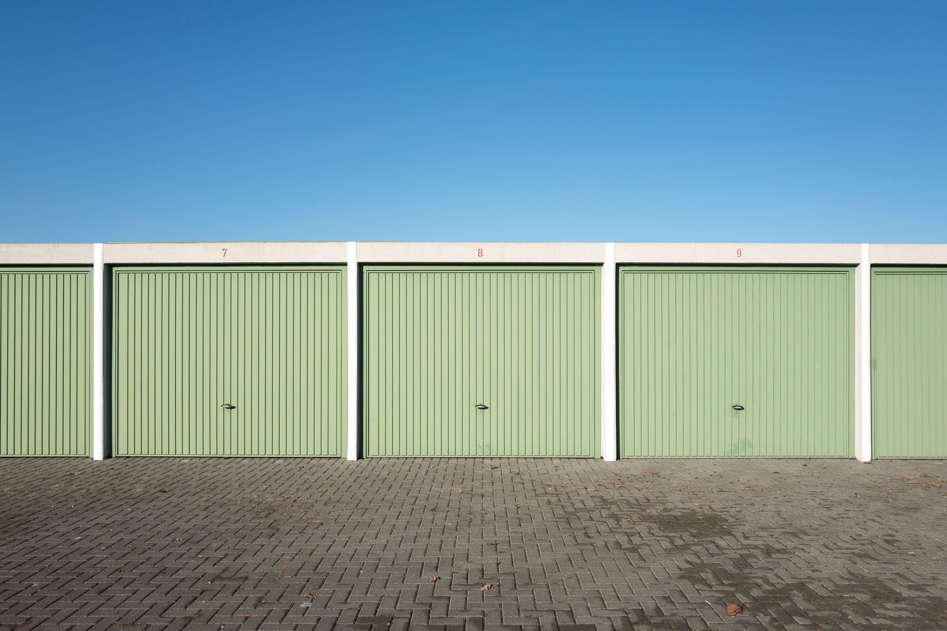 A row of closed, light green metal garage doors with white pillars under a clear blue sky.