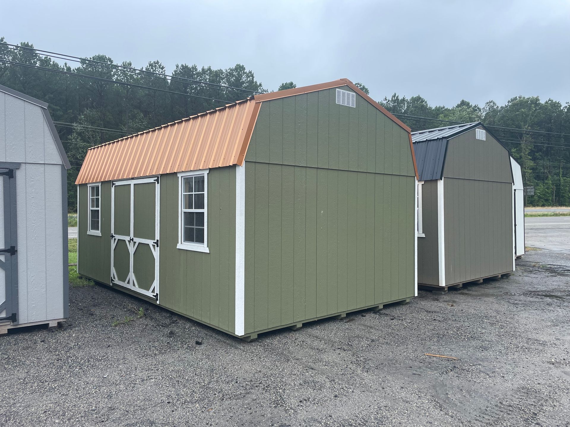 A row of three outdoor storage sheds, with a prominent sage green barn-style shed featuring an orange metal roof.