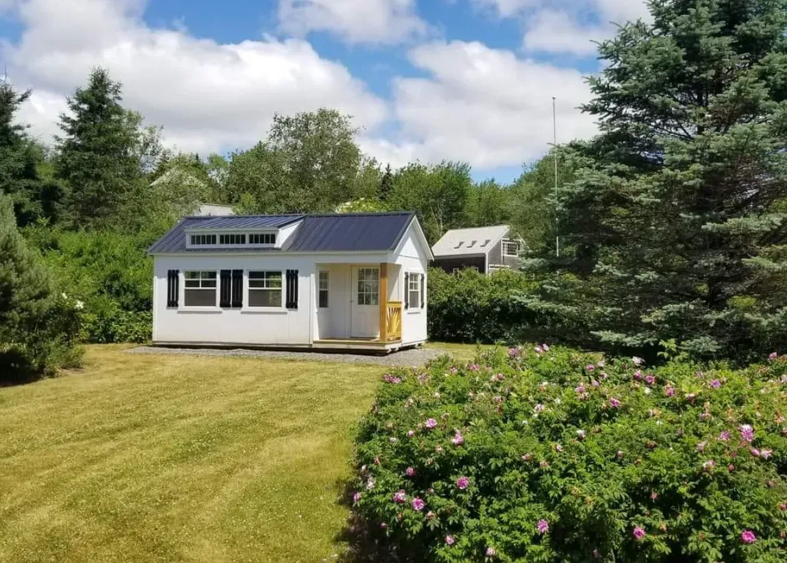 A white, modern tiny house with a dark roof sits on a green lawn surrounded by trees and flowering bushes.