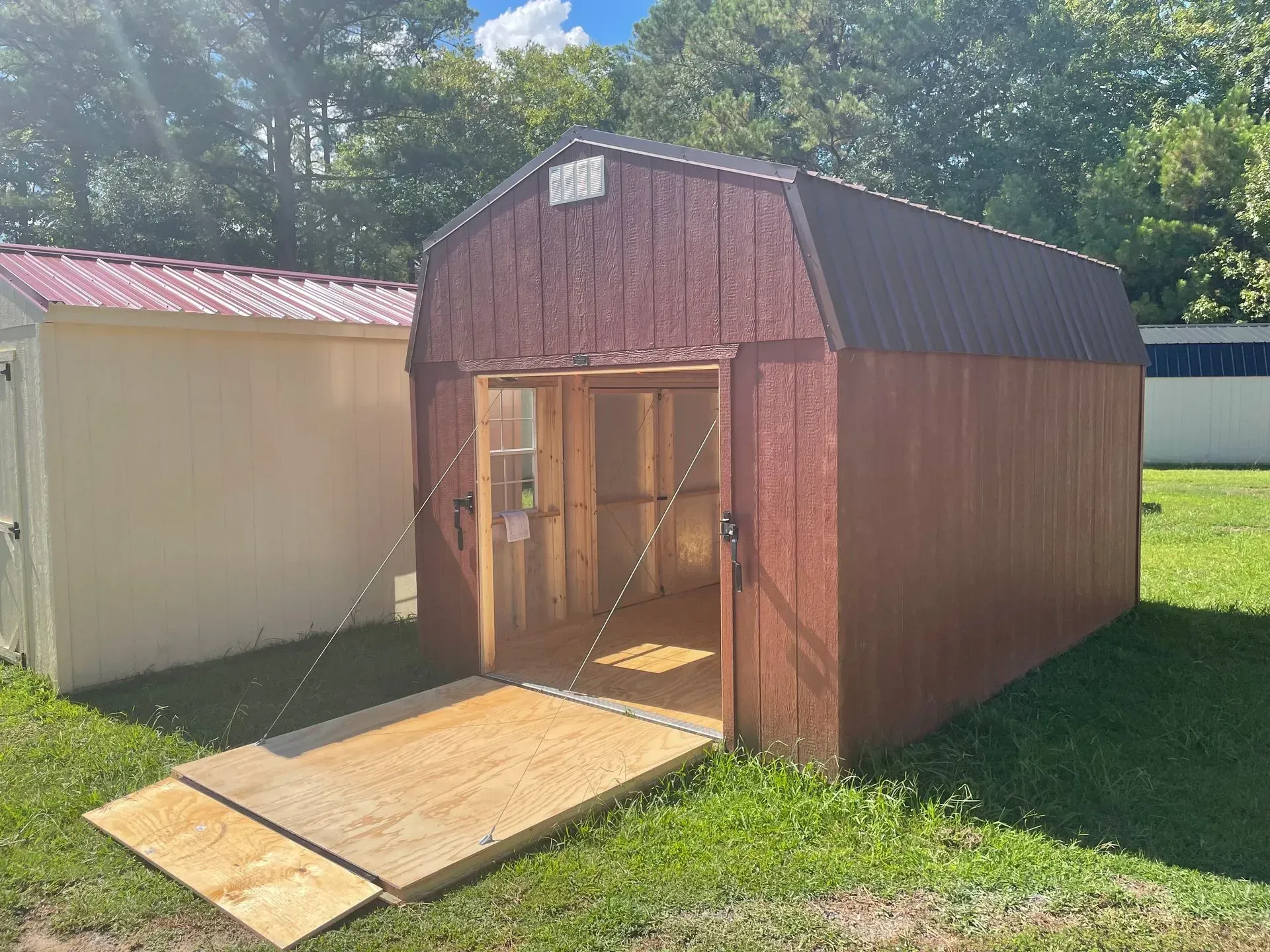 A red-brown wooden storage shed with a barn-style roof and an open ramp door, situated on a grassy lot.