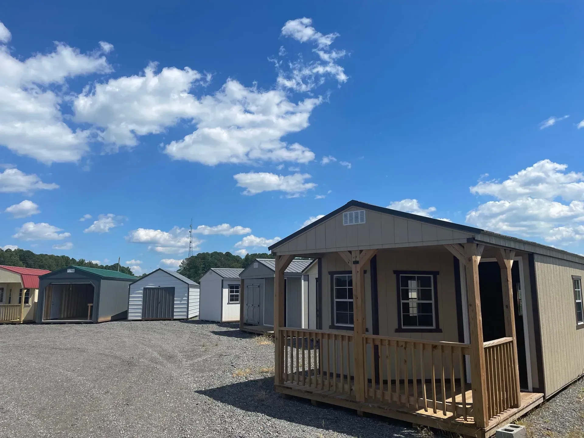 A display lot featuring several small wooden storage sheds and cabins under a bright blue sky with scattered clouds.