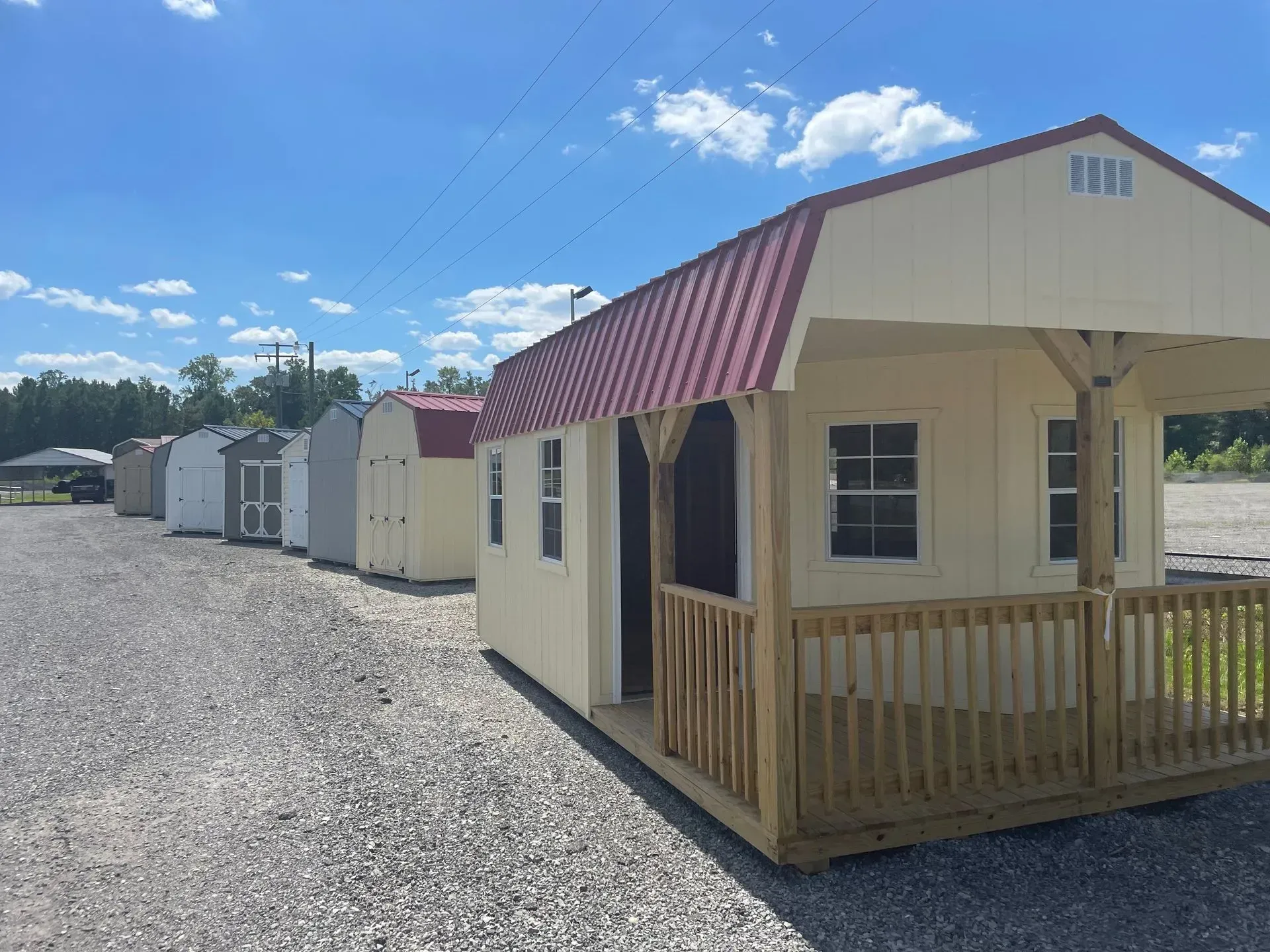 A row of various small, prefabricated portable sheds and cabins parked in a gravel lot under a bright, sunny blue sky.