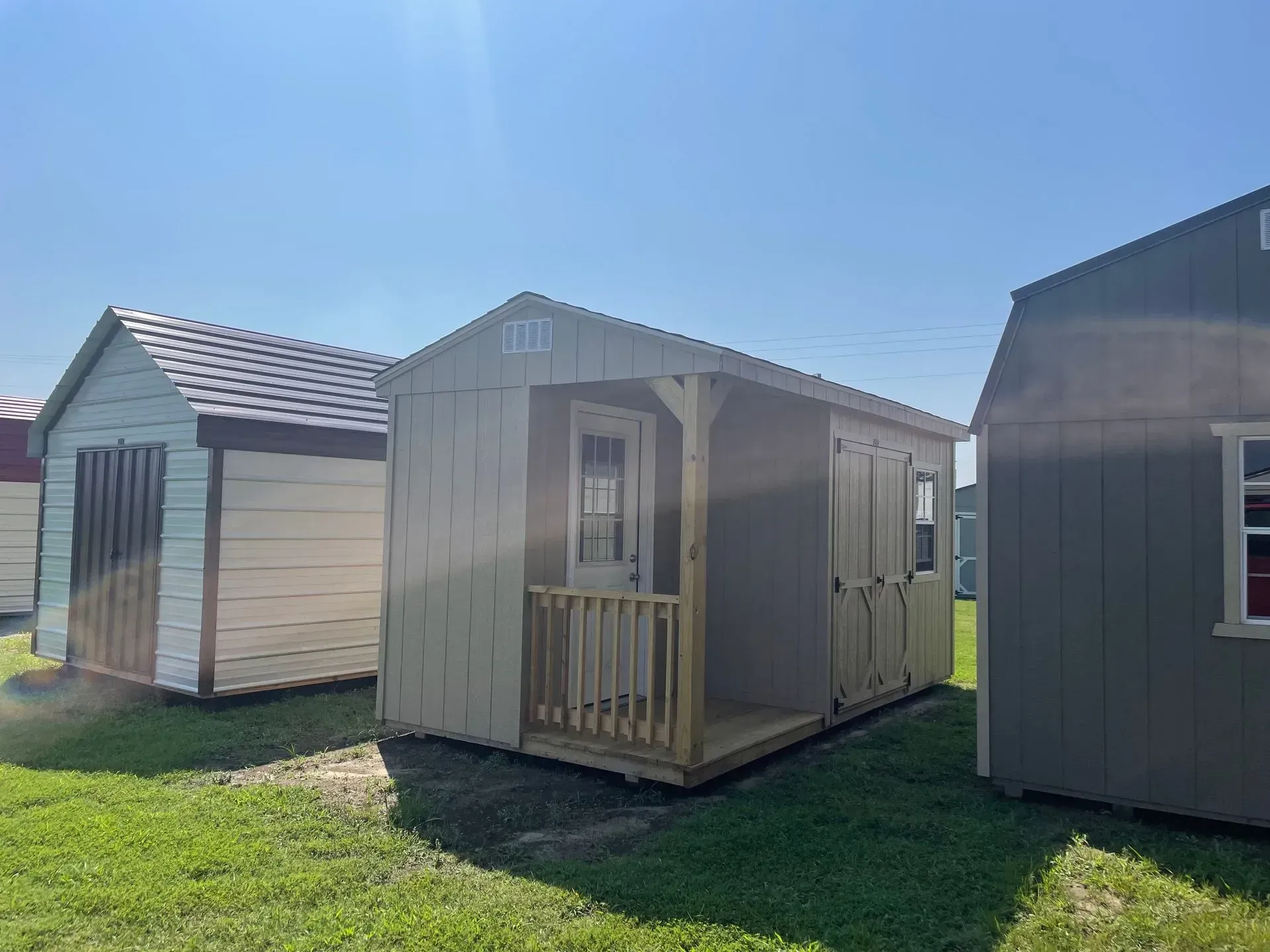 Three outdoor storage sheds displayed on grass under a clear blue sky, including one with a small porch and entry door.