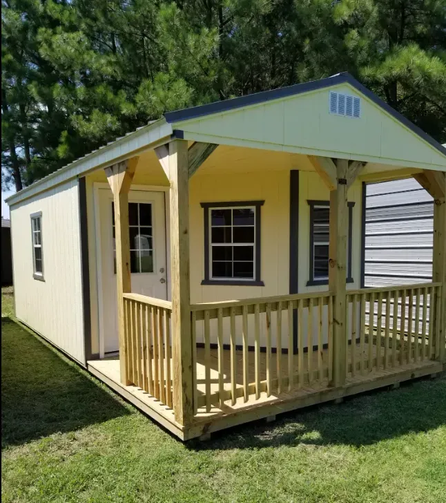 A light yellow wooden shed with a covered front porch, dark trim, and windows, set on a grass lawn near trees.