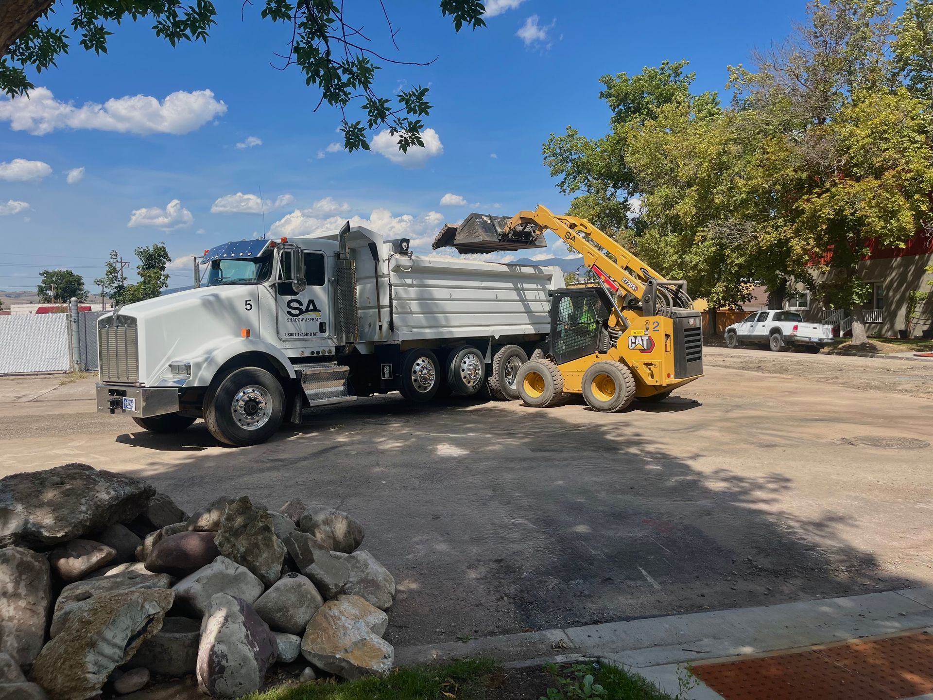 White dump truck being loaded by yellow skid steer in a paved area, under a sunny sky.