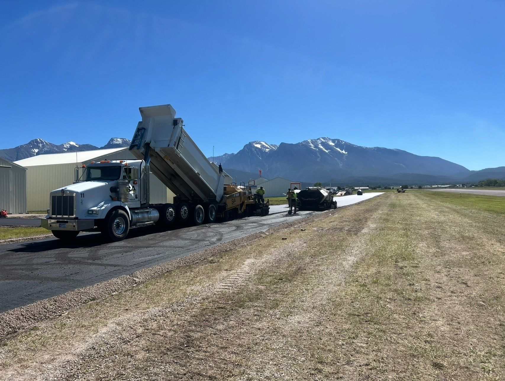 Asphalt paving with dump truck at airfield, mountains in background.