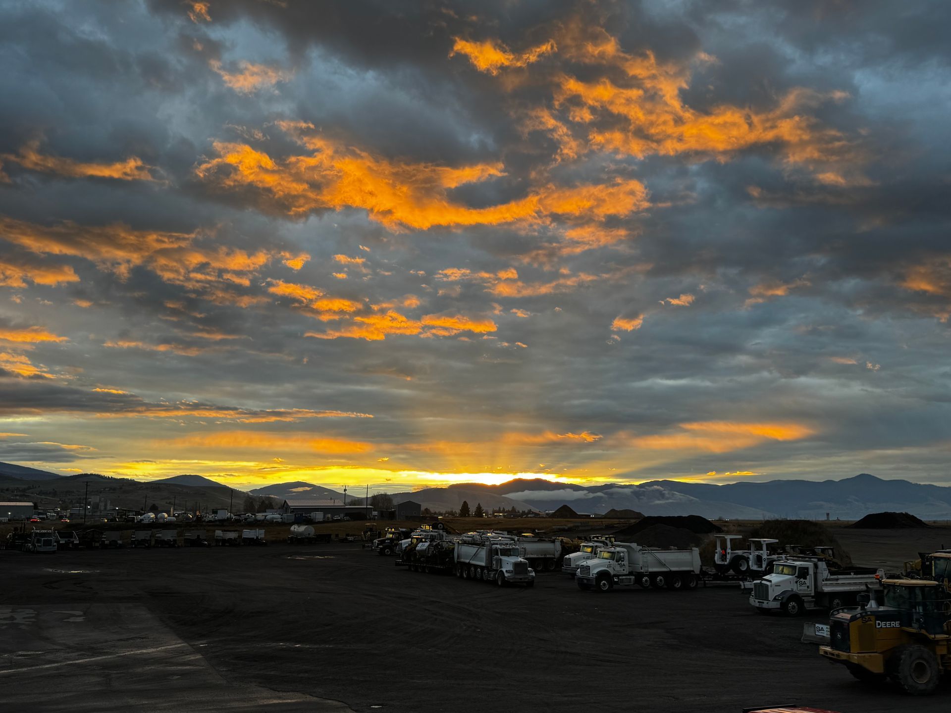 Sunset over a fleet of dump trucks parked on a lot. Orange and yellow clouds illuminate the sky.