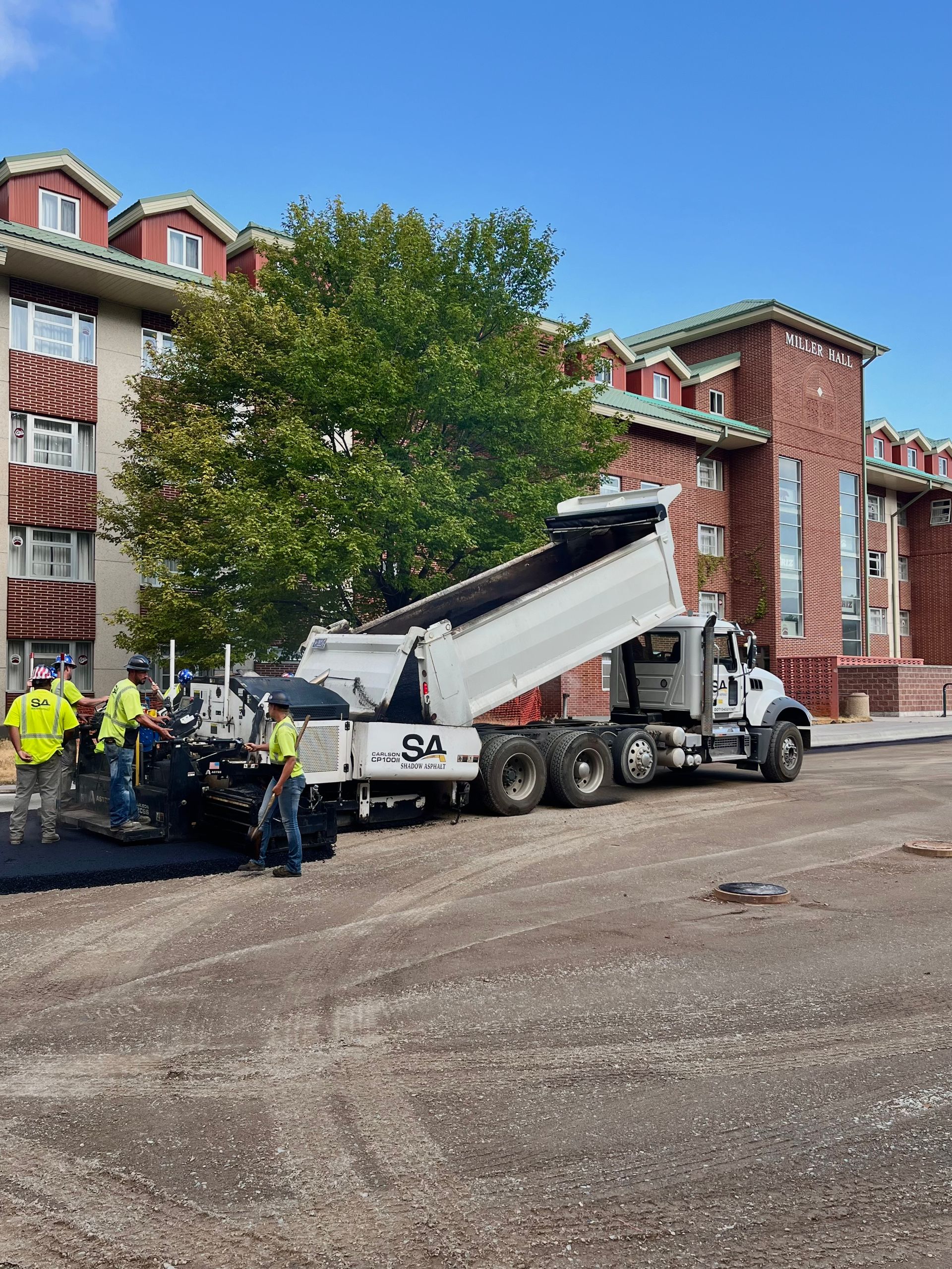 Asphalt paving project: Truck dumping asphalt into a paving machine near a brick building; construction workers present.