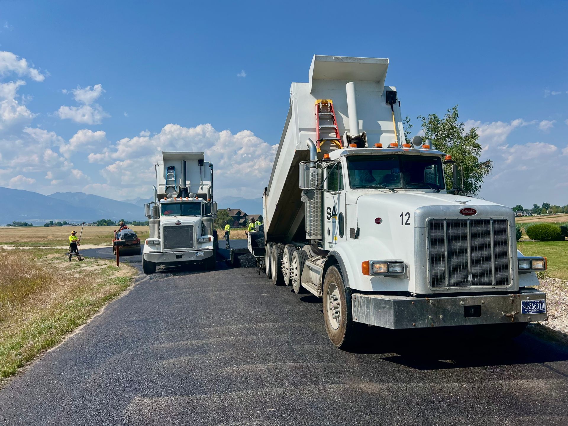 Two white dump trucks unloading asphalt on a newly paved road, with workers in the background. Blue sky and mountains.