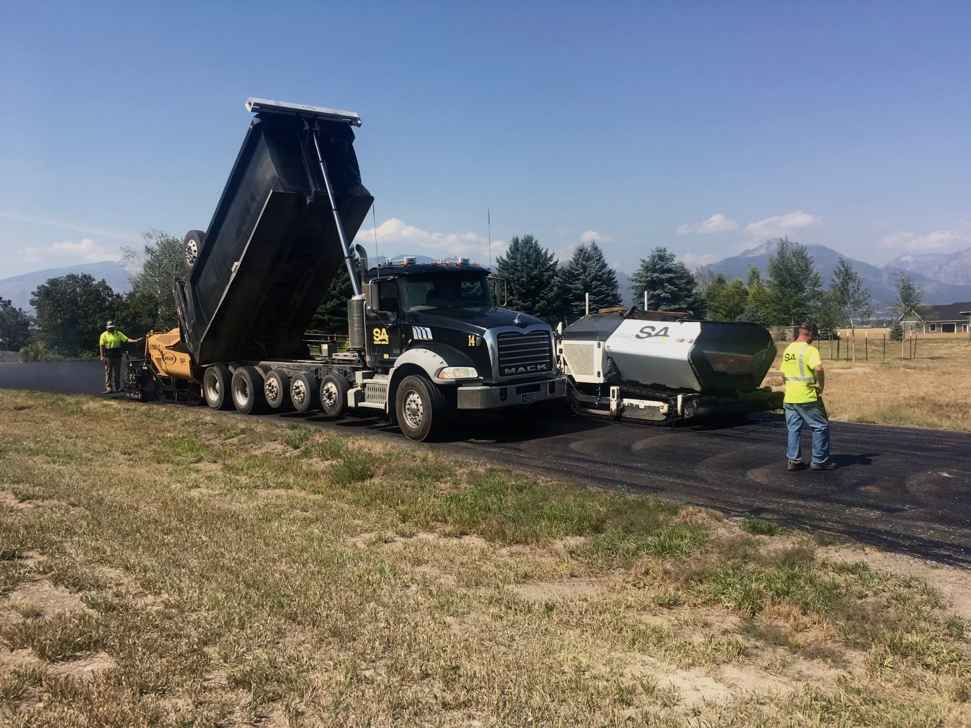 Asphalt paving in progress: Dump truck unloading black asphalt into a paver on a sunny day. Two workers.