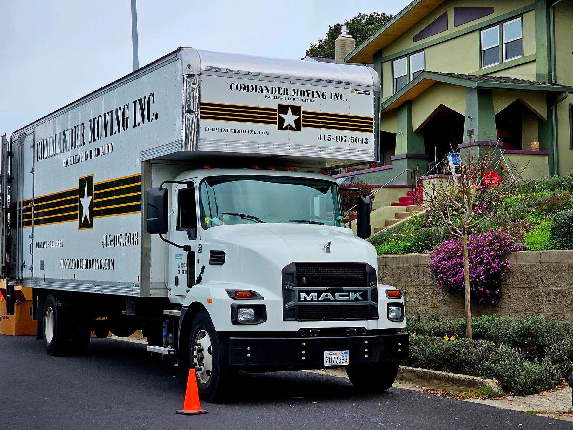 White Mack truck with moving company branding parked in front of a green house. Orange cone present.