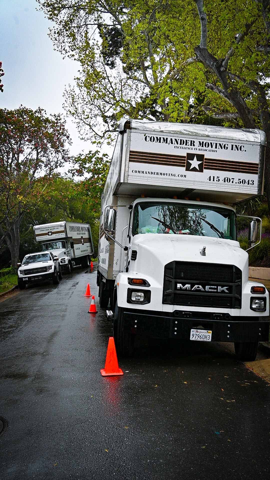 White Mack truck and two moving trucks on a wet street, orange cones, car, trees. 
