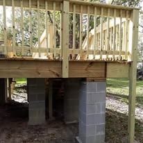 A newly constructed wooden deck featuring vertical railings, supported by several concrete block pillars on a dirt yard.