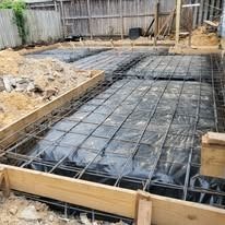 A concrete slab foundation under construction, featuring a metal rebar grid laid over a black plastic moisture barrier.