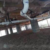 White PVC plumbing pipes hang from wooden floor joists above a dirt crawl space with concrete block support piers.