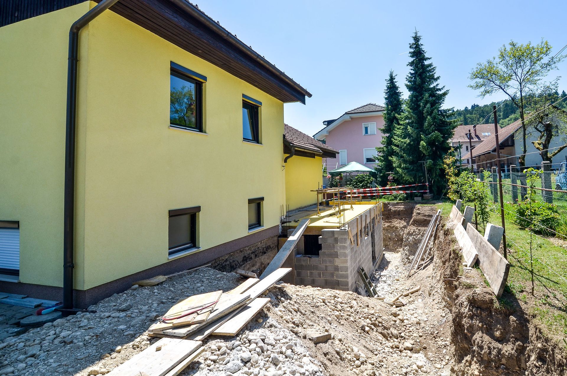 A yellow house under construction with an excavated foundation and gravel ground in a sunny outdoor setting.