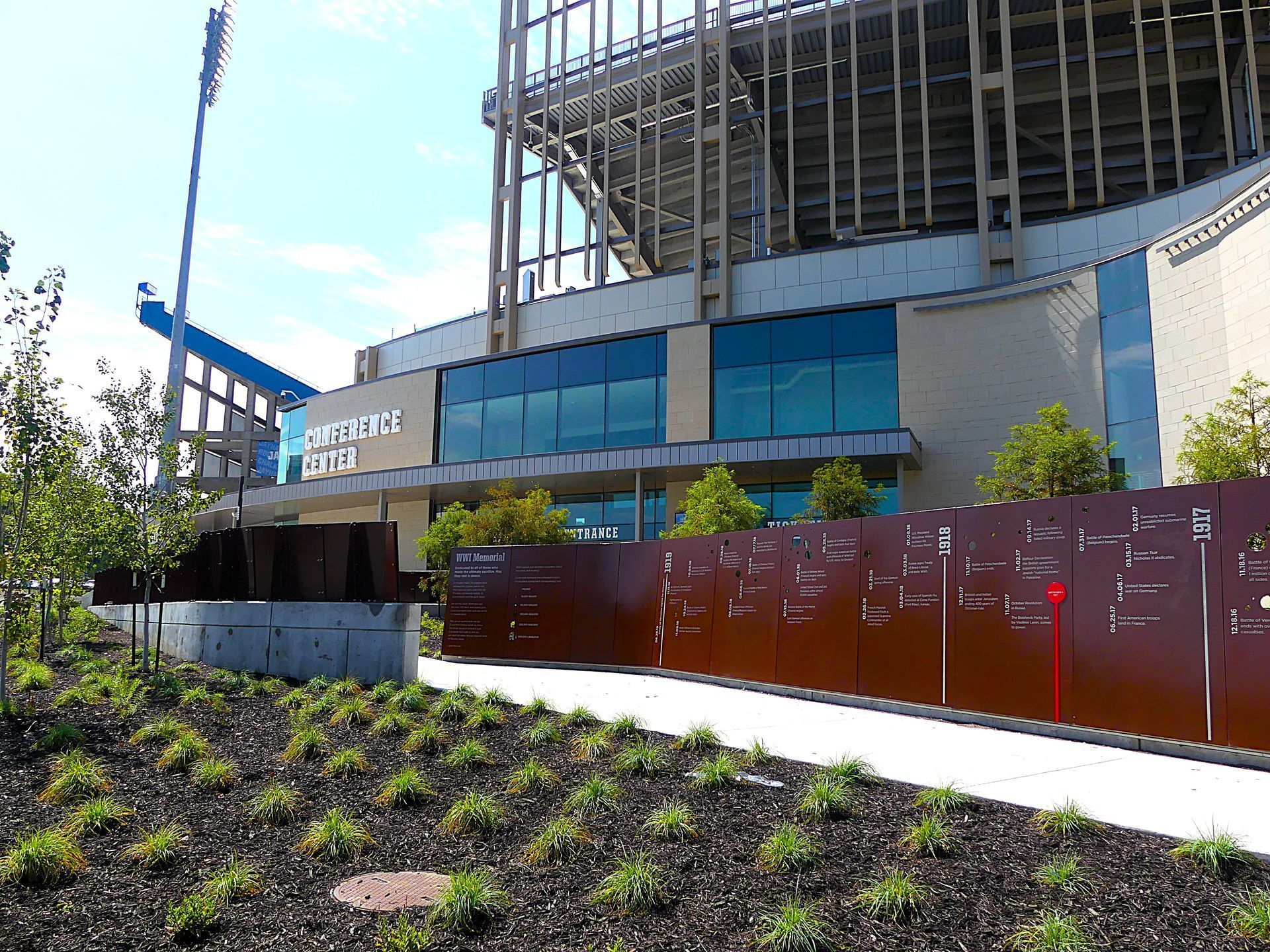 Exterior of a stadium with a brown metal fence, landscaping, and clear blue sky.