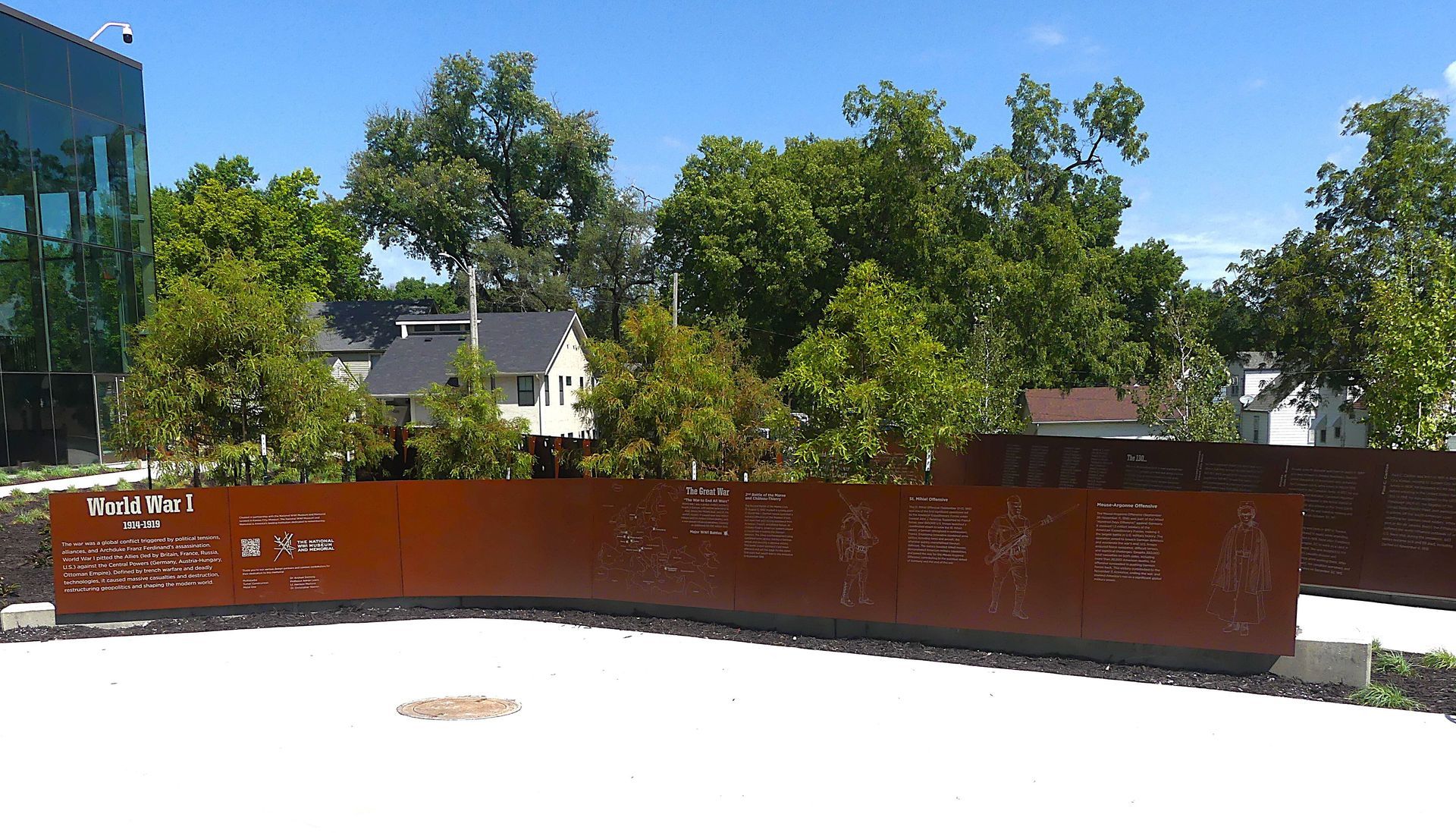 Rust-colored metal wall with etched text, trees, and a modern building on a sunny day.