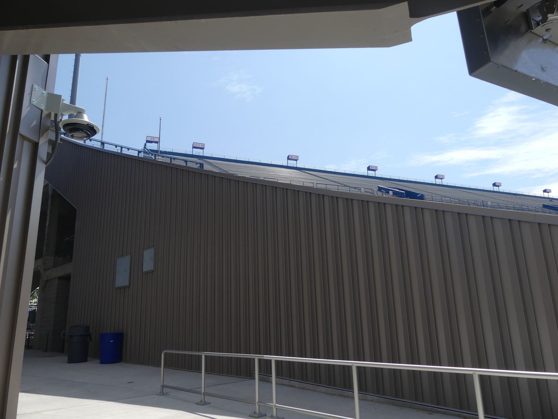 Side view of a light brown, corrugated building under a clear blue sky, with a walkway and security camera.