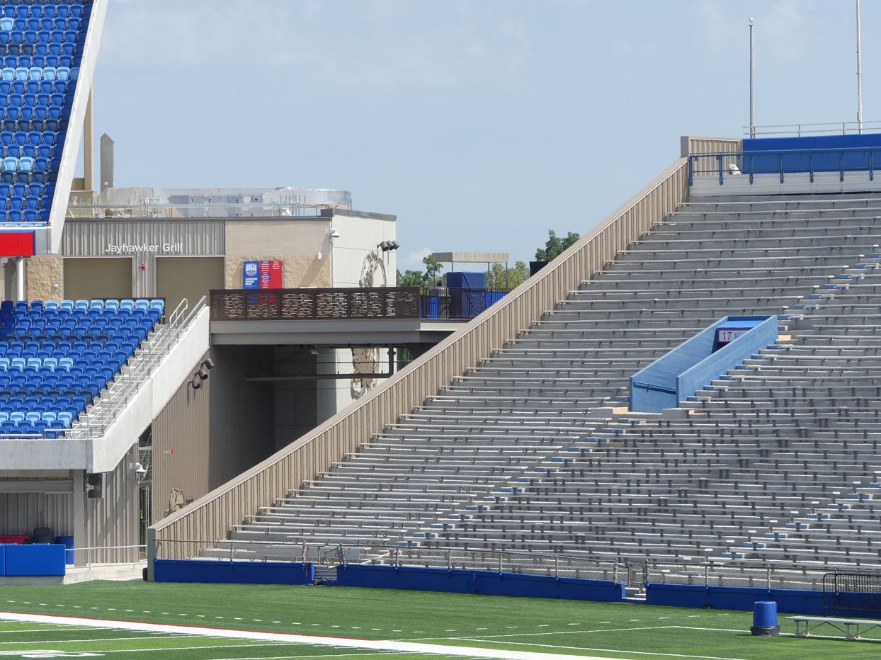 Empty stadium seats, blue and gray, with a walkway and service building visible in daylight.