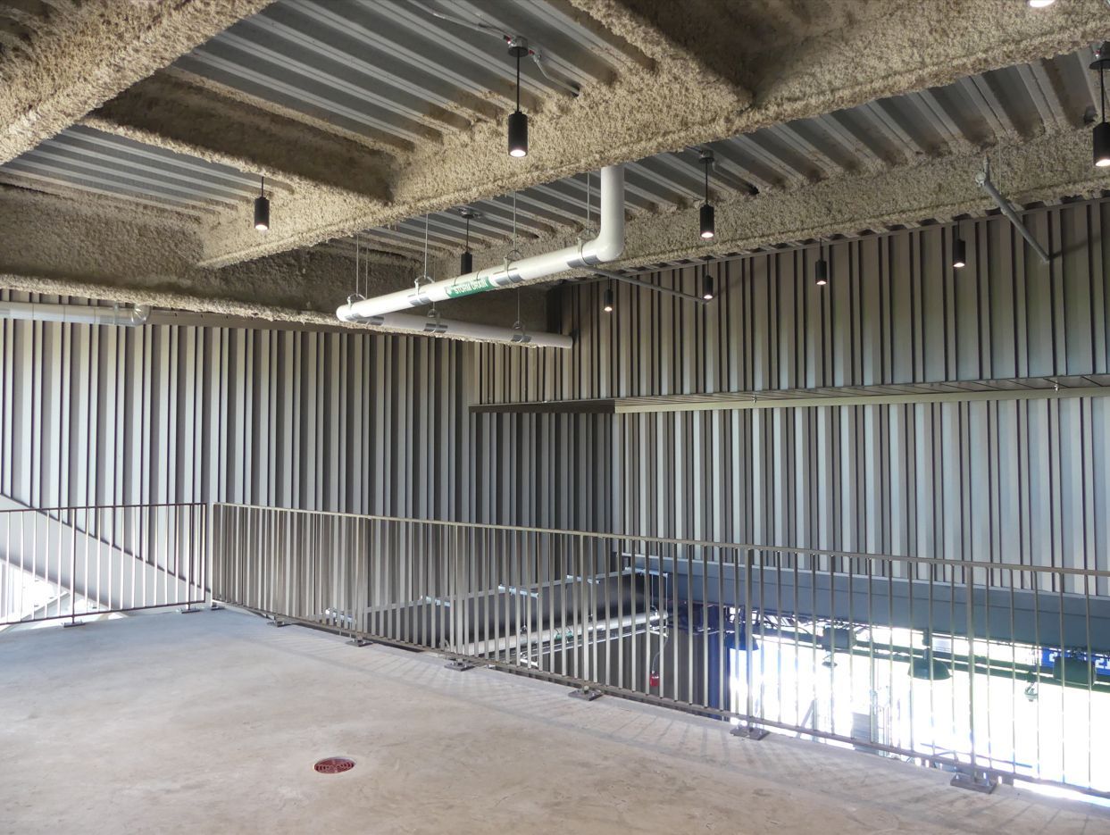 Interior view of unfinished building with metal beams and ceiling, exposed piping, and railings.